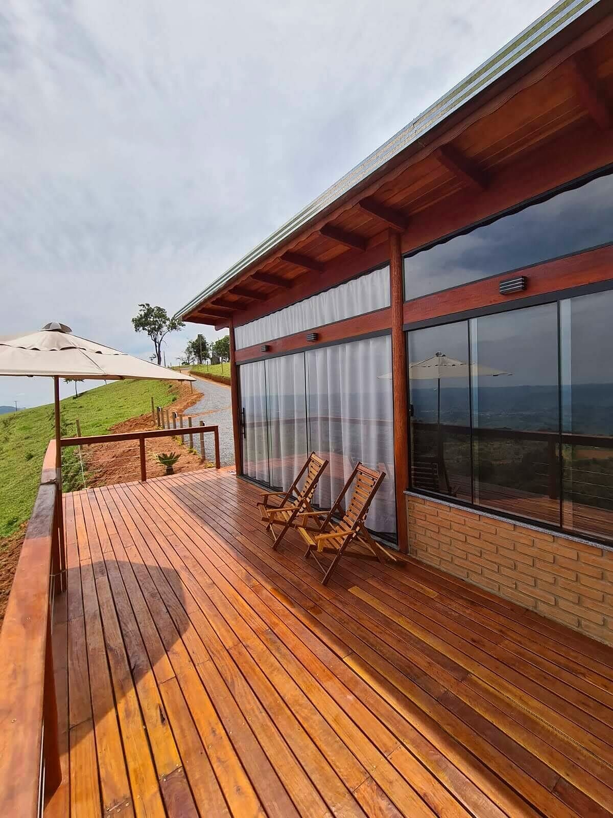 Deck de madeira envernizada do chalé com duas espreguiçadeiras e guarda-sol, de frente para morros verdes