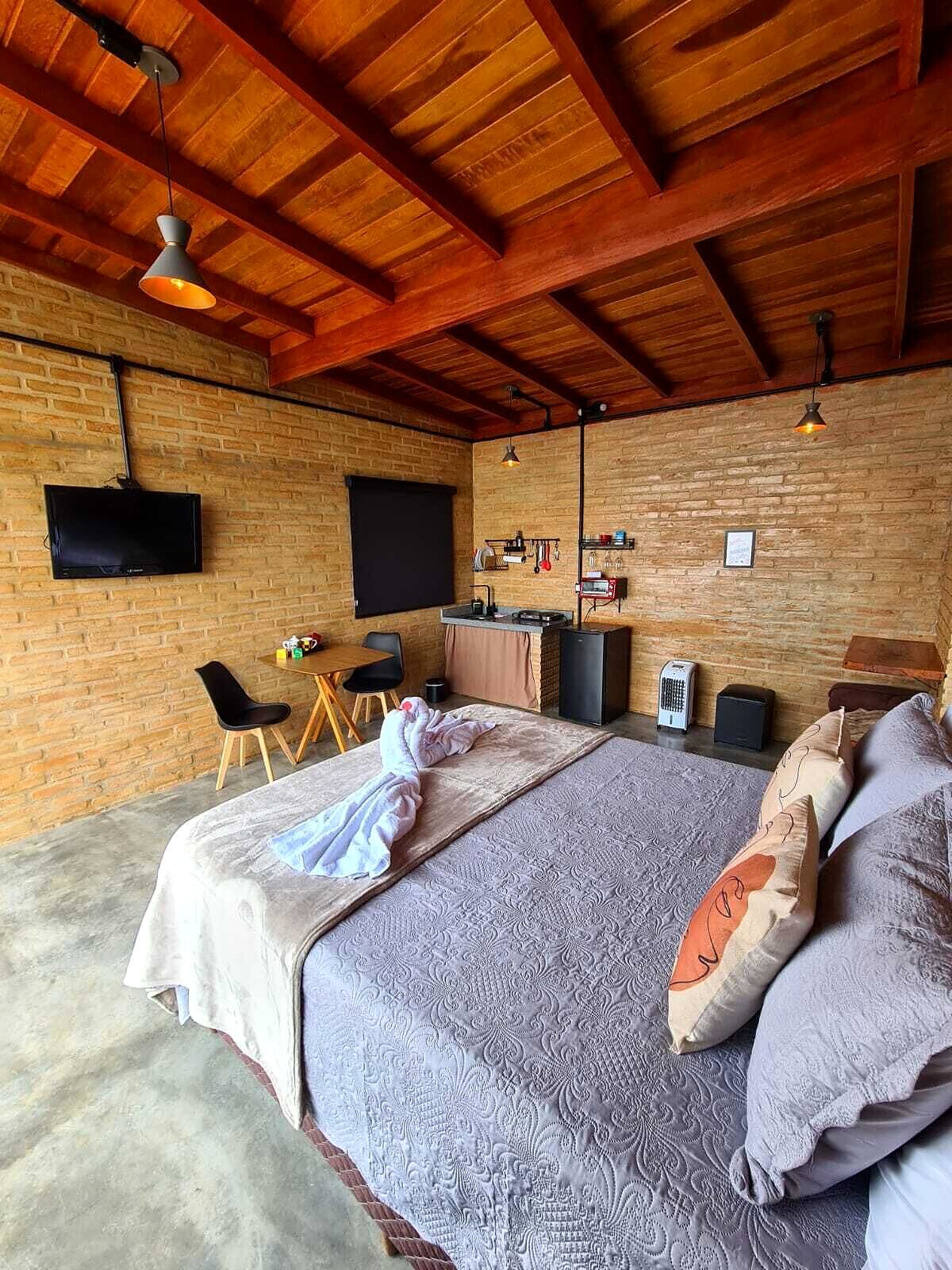 Interior do chalé com cama de casal, paredes de tijolo aparente, forro de madeira com vigas e cozinha compacta ao fundo