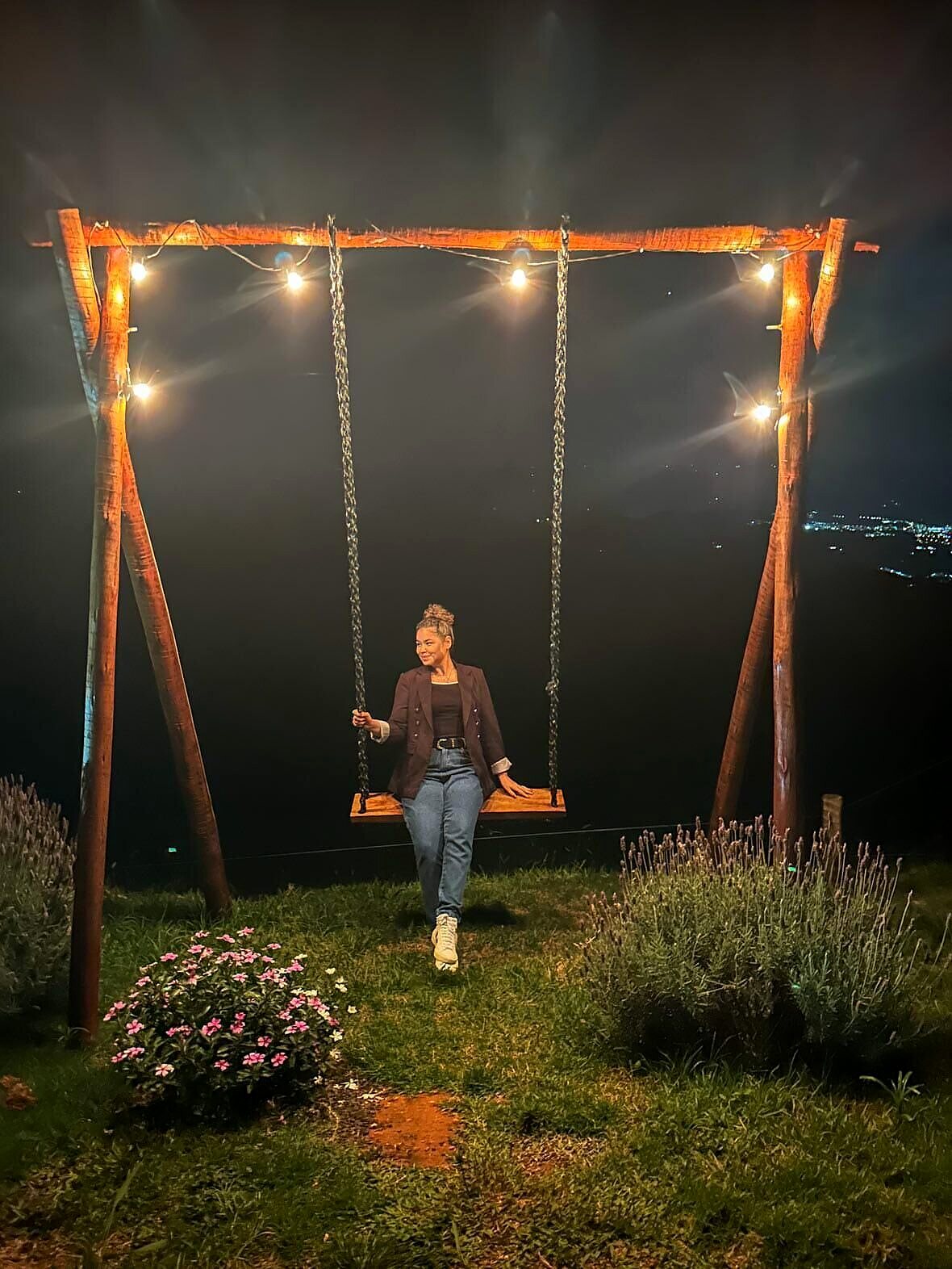Balanço de madeira iluminado por varal de luzes à noite, com lavandas ao redor e as luzes da cidade brilhando no vale lá embaixo